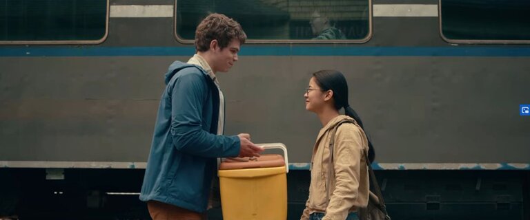 Paul and Ellie stand facing each other next to the train on which Ellie is to depart. Paul is holding a large cooler containing food for Ellie’s journey.