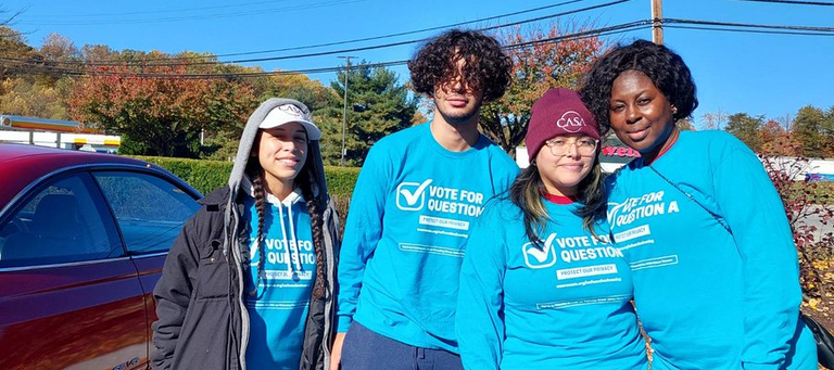 A photograph of three young women and one young man. They are wearing light blue shirts printed with the slogan “Vote for Question A.”